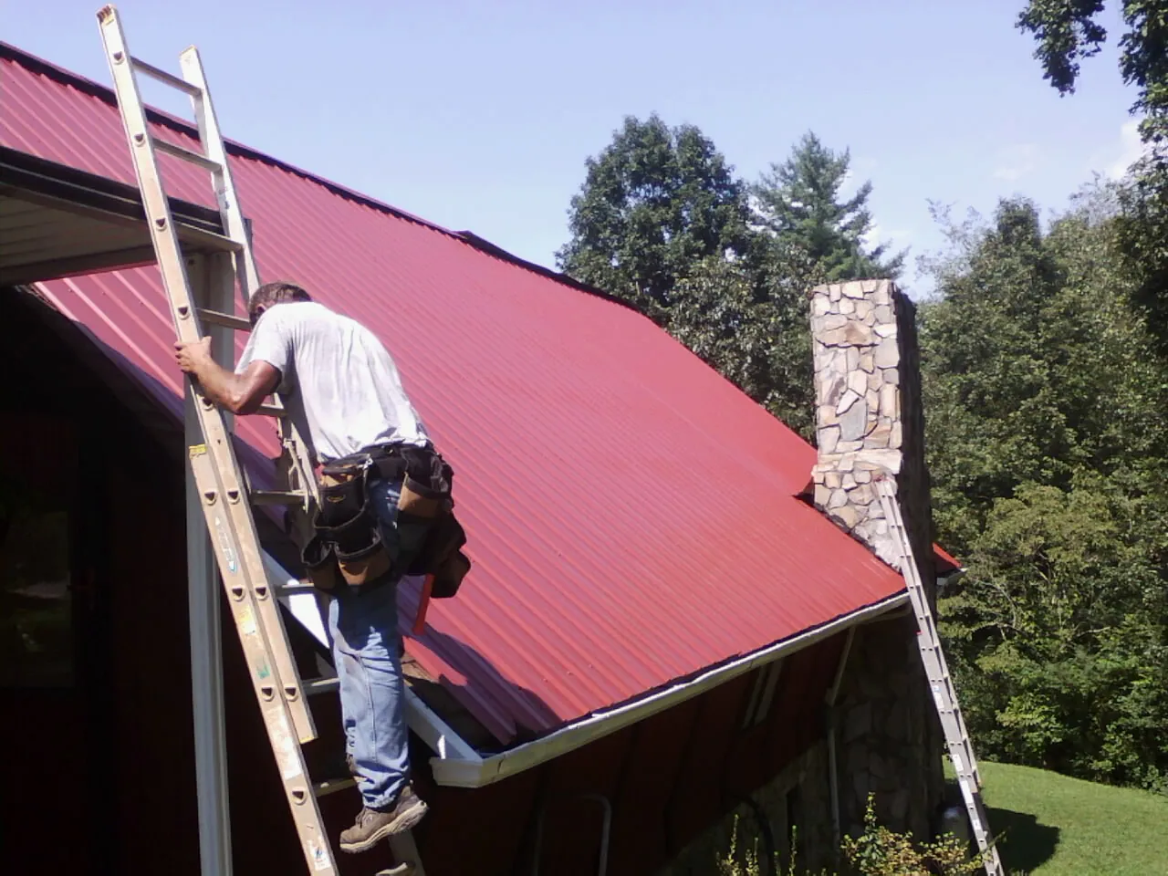 Roofer on a ladder beside a red metal roof and stone chimney — Soffit & Fascia Repair by Roofing Brothers in Awendaw